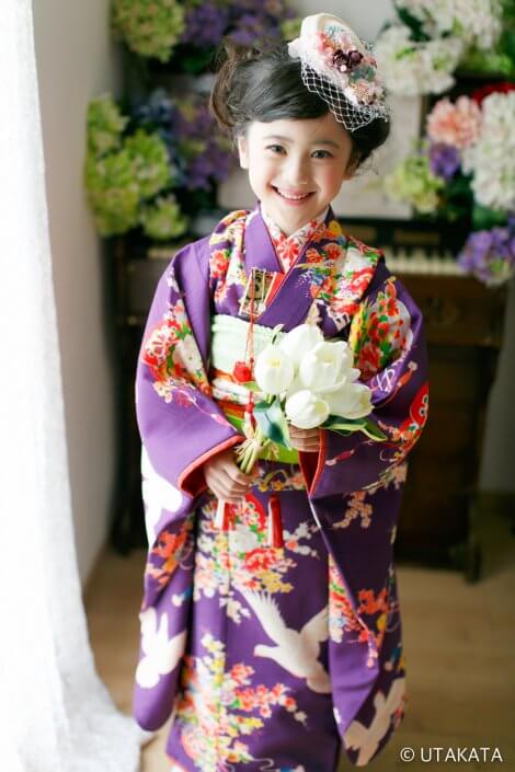 Young girl in a purple floral kimono, holding a white bouquet and smiling for a ceremony.