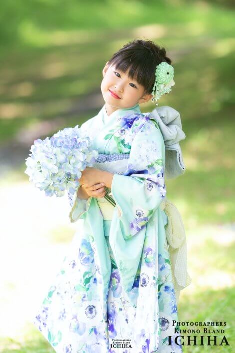 Young girl in a mint and lavender kimono holding a blue bouquet, standing in a sunlit park and smiling at the camera.