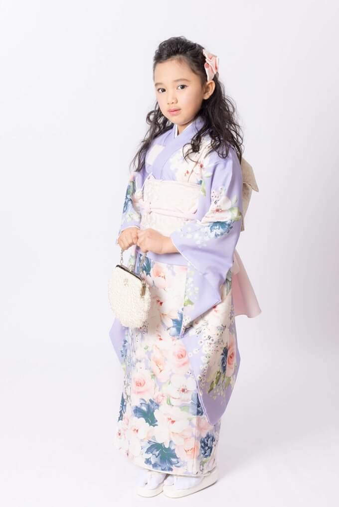 Young girl in a lavender floral kimono with a pale pink obi, holding a small white handbag against a white backdrop.