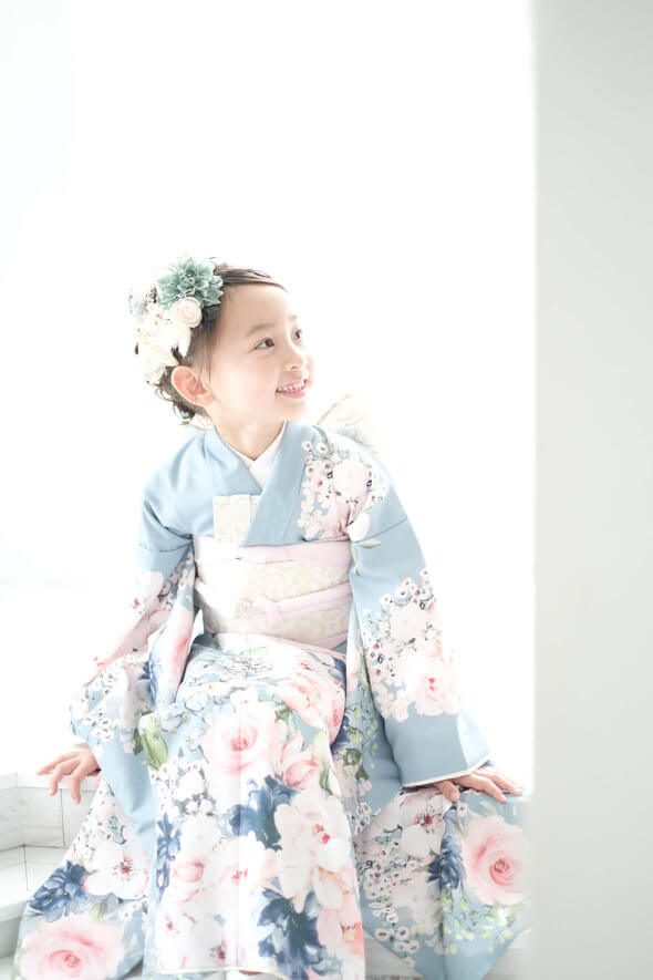 Young girl in a light blue floral kimono sitting by a bright window, smiling softly.