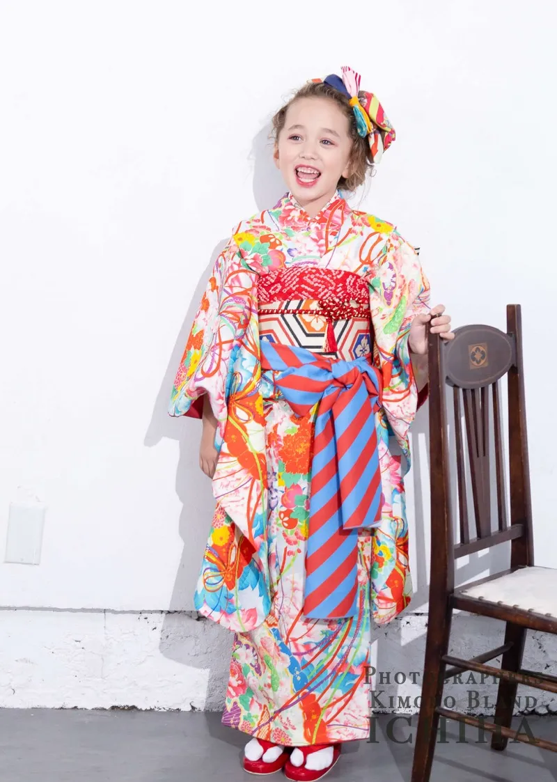 Girl in a vibrant floral kimono with red obi, standing beside a wooden chair and smiling joyfully.