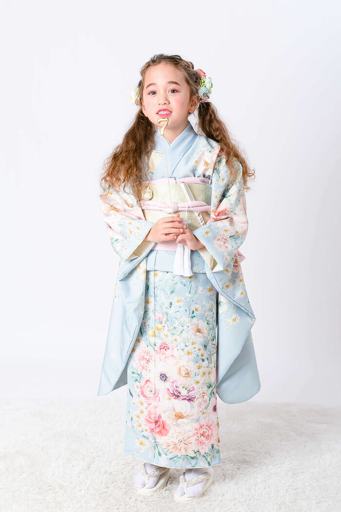 Girl in a pastel blue floral kimono with a pink sash, standing on a white rug with hands clasped and flower hair accessories in a studio setting.