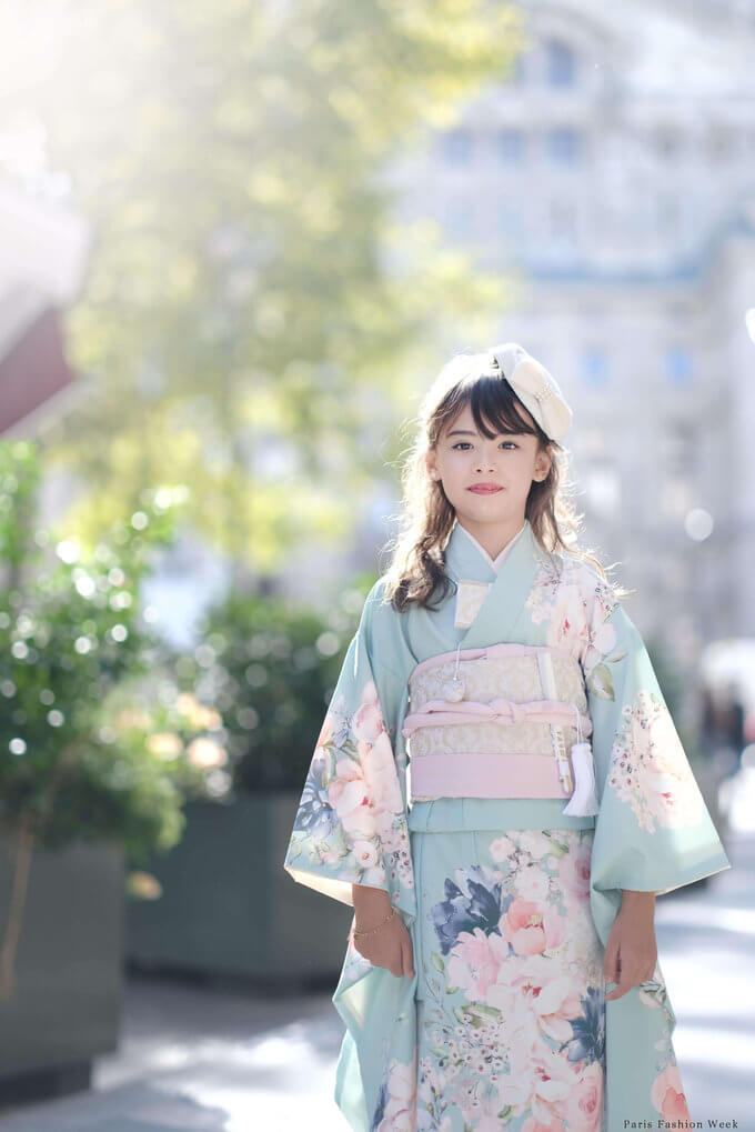 Young girl wearing a light blue floral kimono with a pink obi, standing on a sunlit city sidewalk.